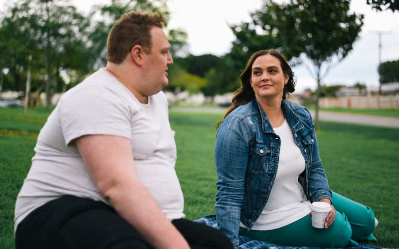 Image from gallery: a man and woman with obesity talk together outside at a park while sitting down on the grass